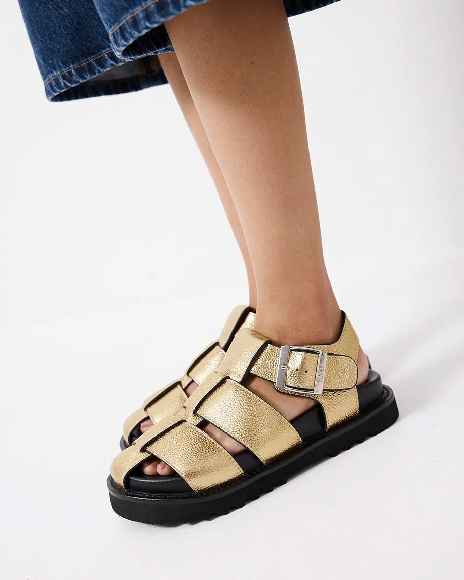 Gold sandals with black soles worn by a person on a white background