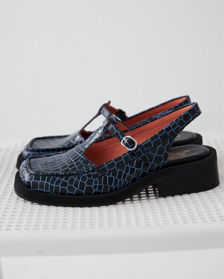 A pair of women's slingback flats in blue croc leather with a silver buckle are placed on a white chair.