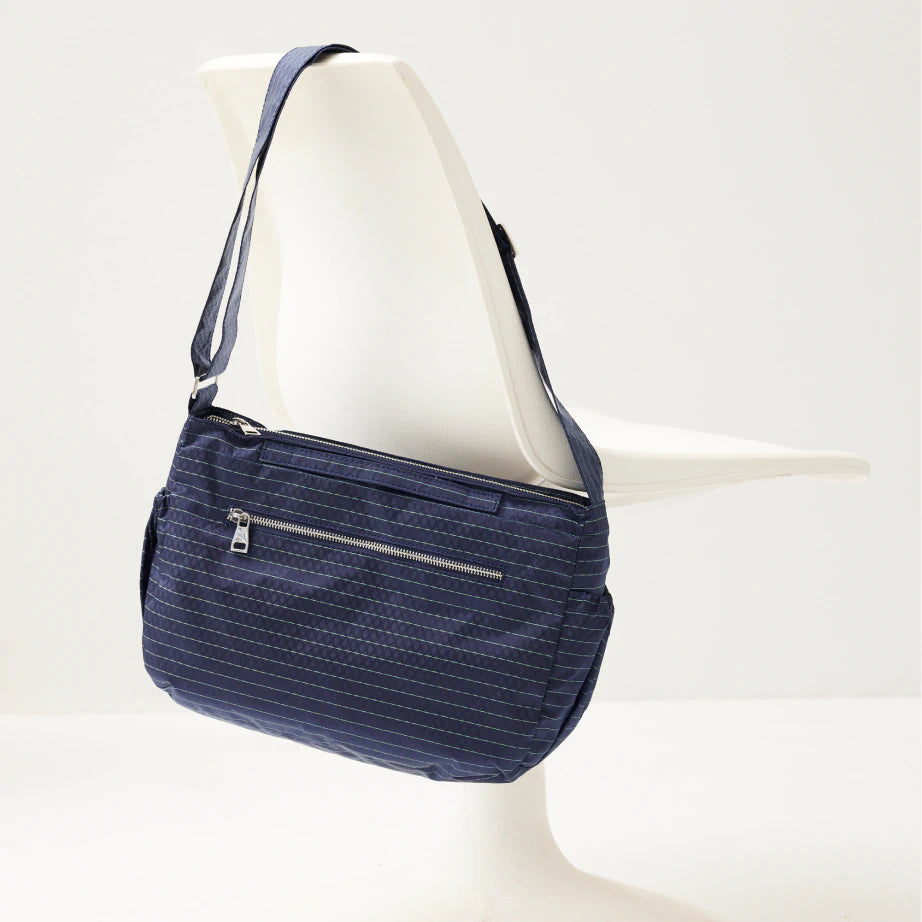 A modern crossbody zip up satchel bag in blue nylon with silver zippers hangs over a modernist tulip chair.