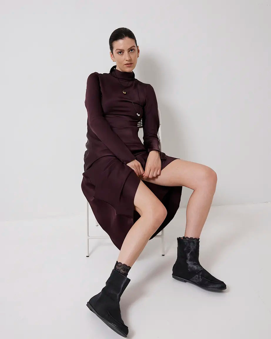 Woman wearing a dark brown dress and black pony hair ankle boots sitting on a white chair against a white background
