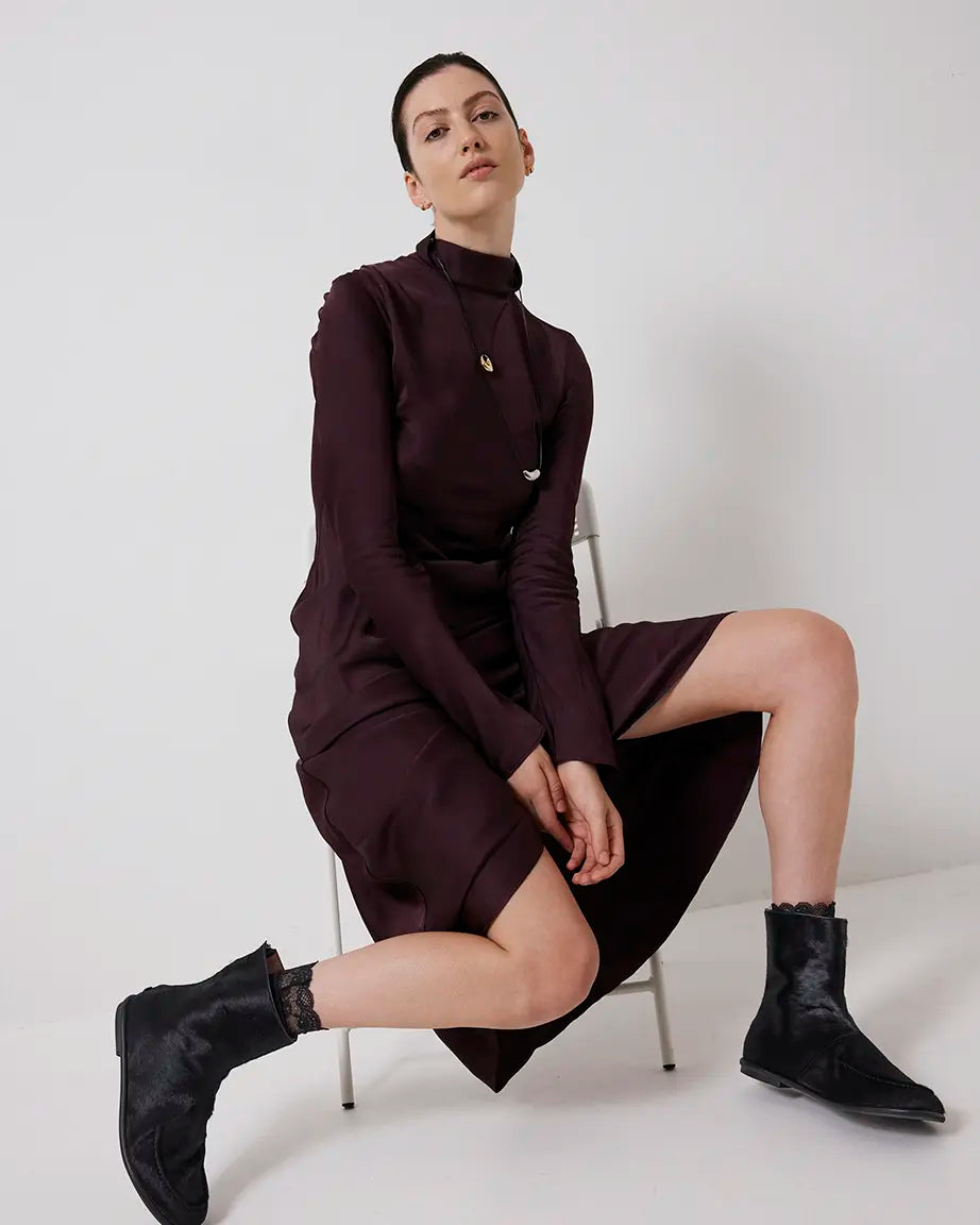 Woman wearing a dark brown outfit with black pony hair ankle boots sitting on a white chair against a white background.