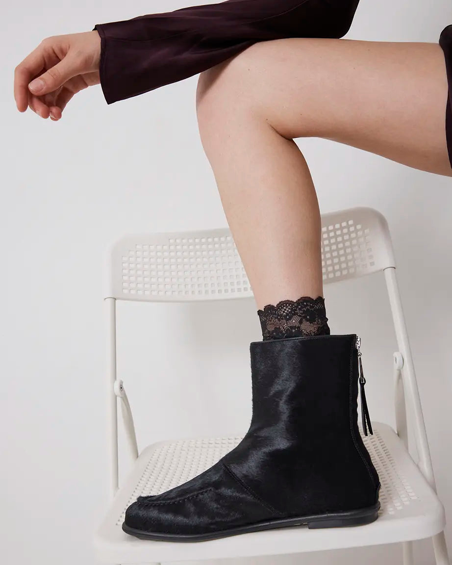 Person wearing black pony hair ankle boots and black lace socks on a white chair.