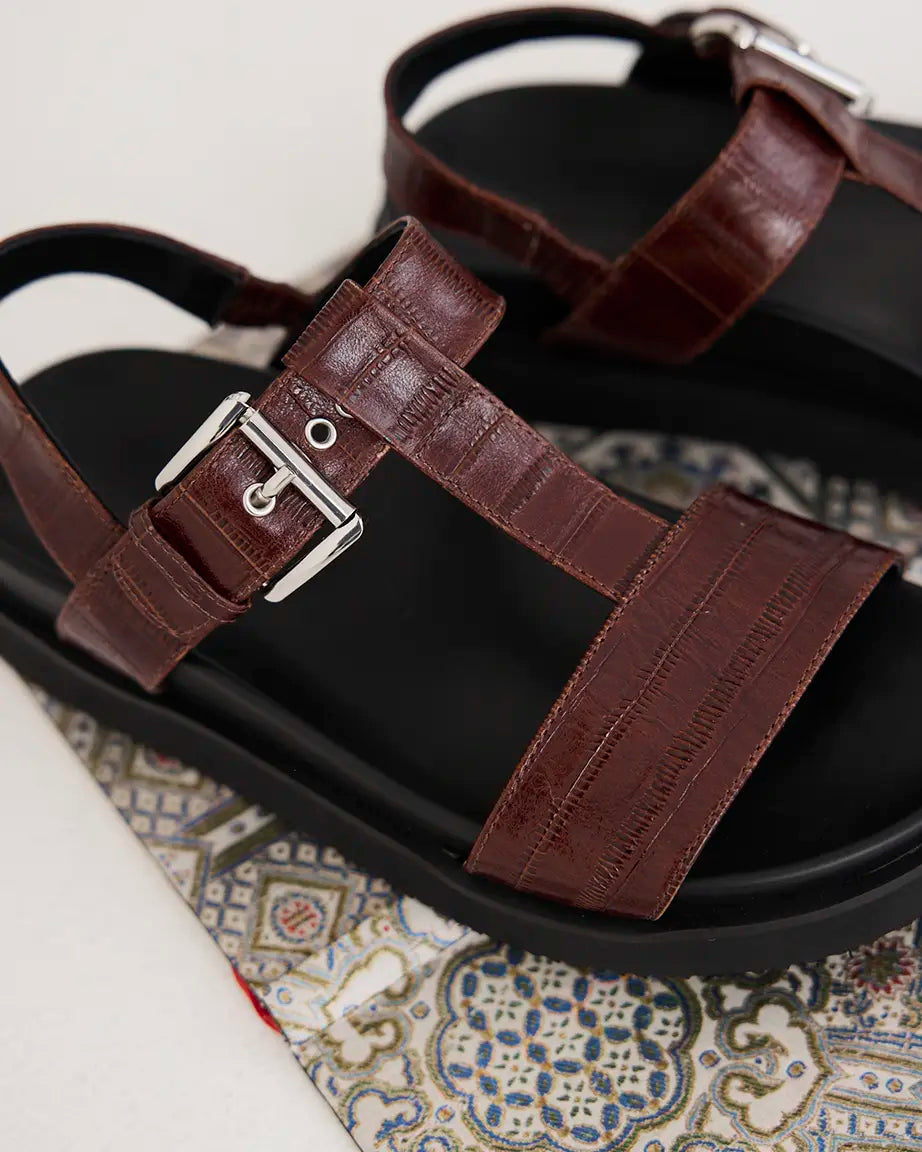 A close-up of dark brown eel-embossed leather strap sandals with silver buckles and black soles.
