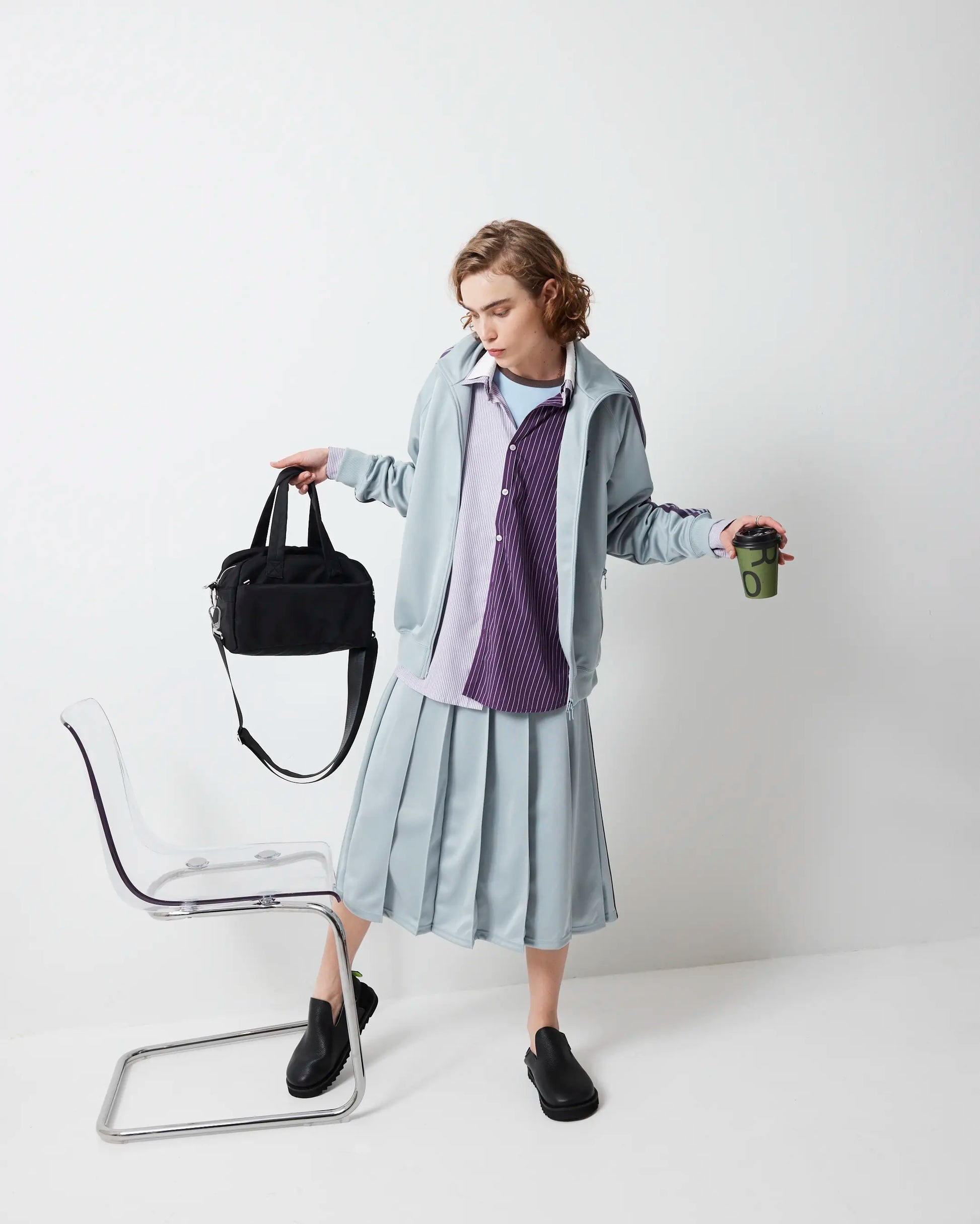 Person wearing a light gray outfit with a purple shirt, holding a black nylon bag