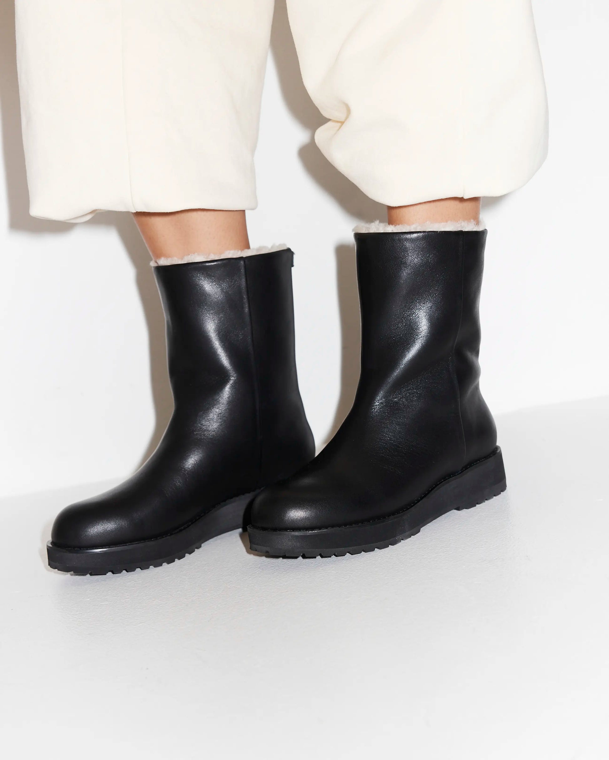Black leather wool lined boots worn with white pants on a light gray background