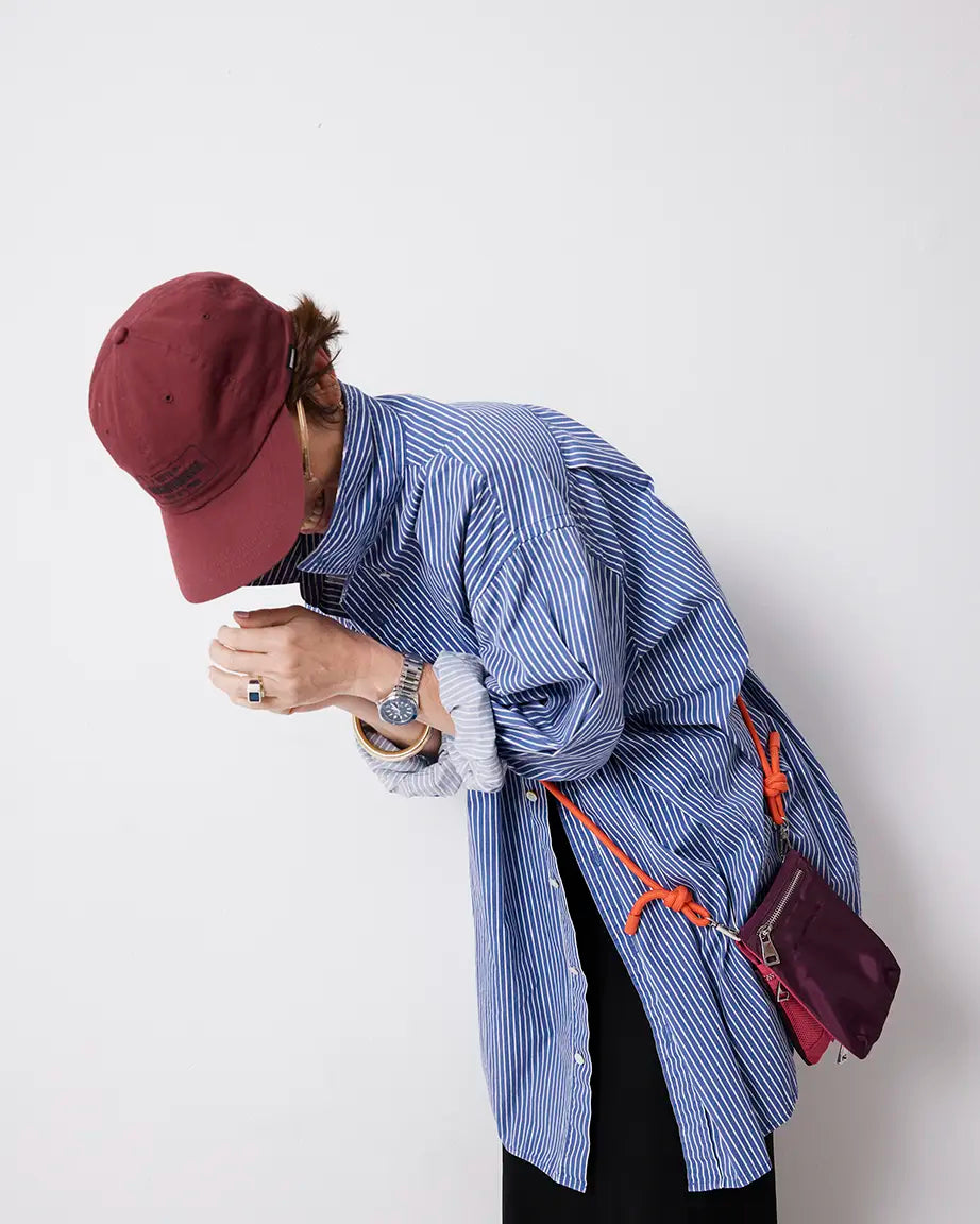 Person wearing a blue striped shirt and red cap, leaning against a white wall wearing a purple phone pouch.