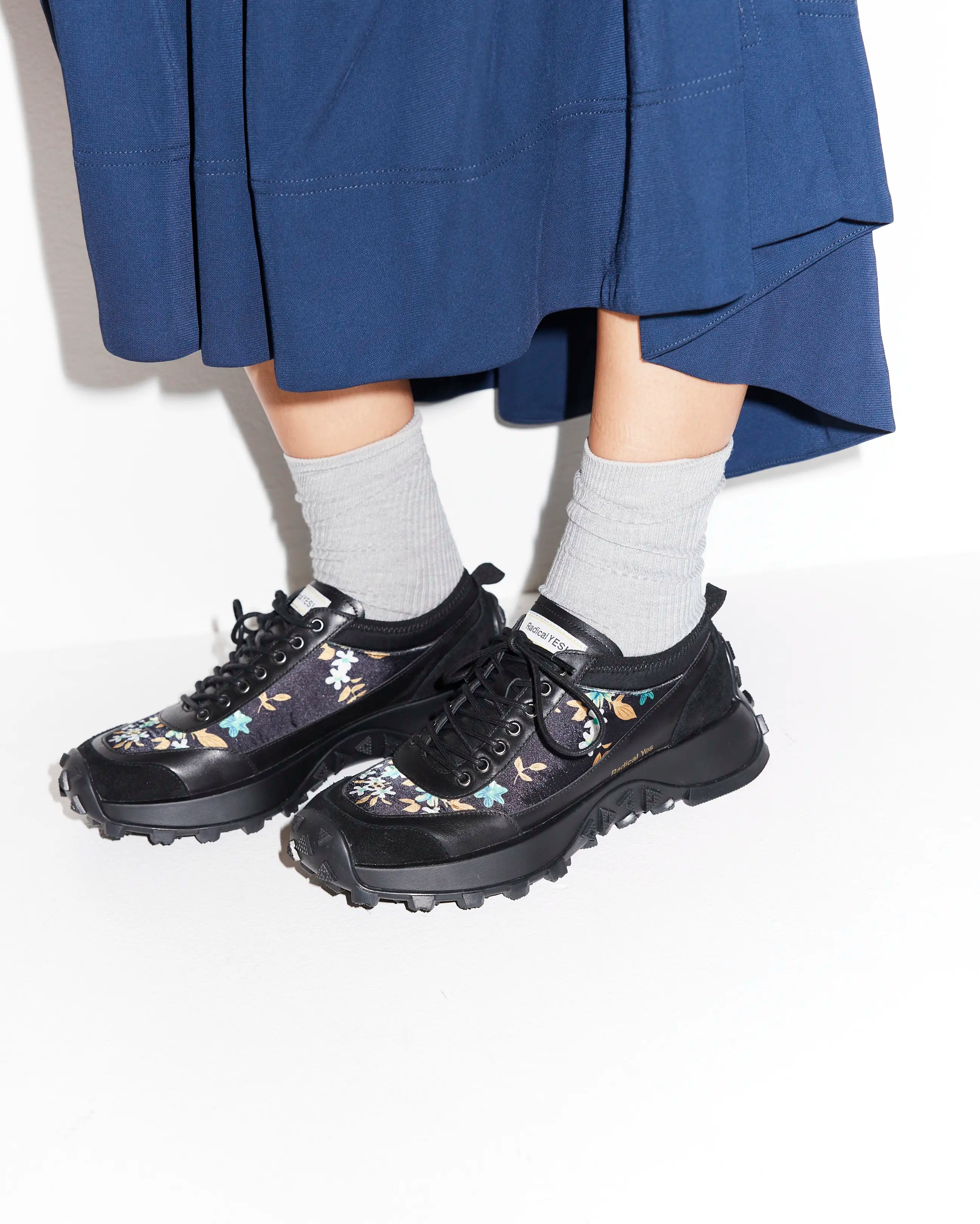 Black floral sneakers worn with gray socks and a blue skirt on a white background