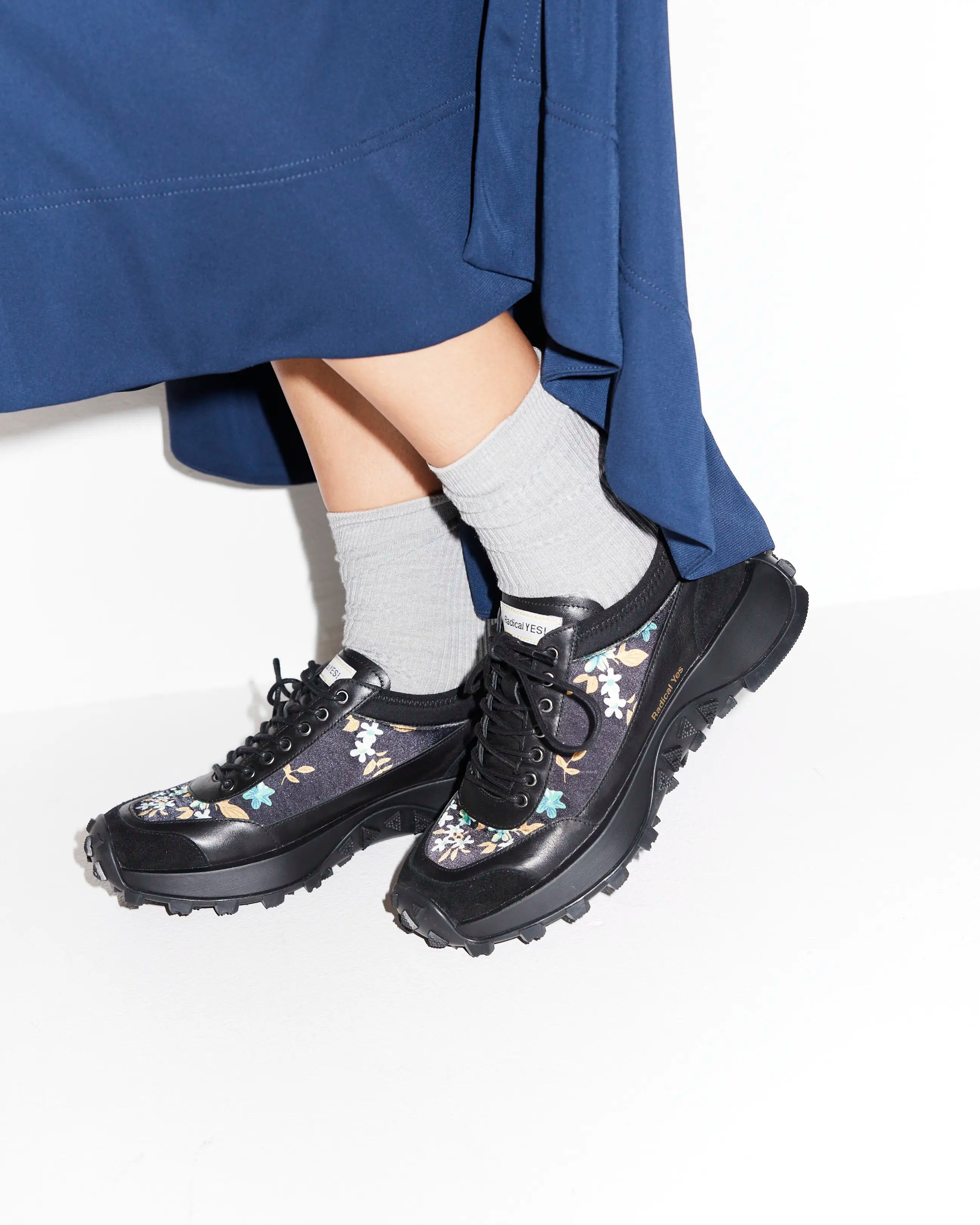 Black floral-patterned shoes worn with a blue skirt and gray socks on a white background