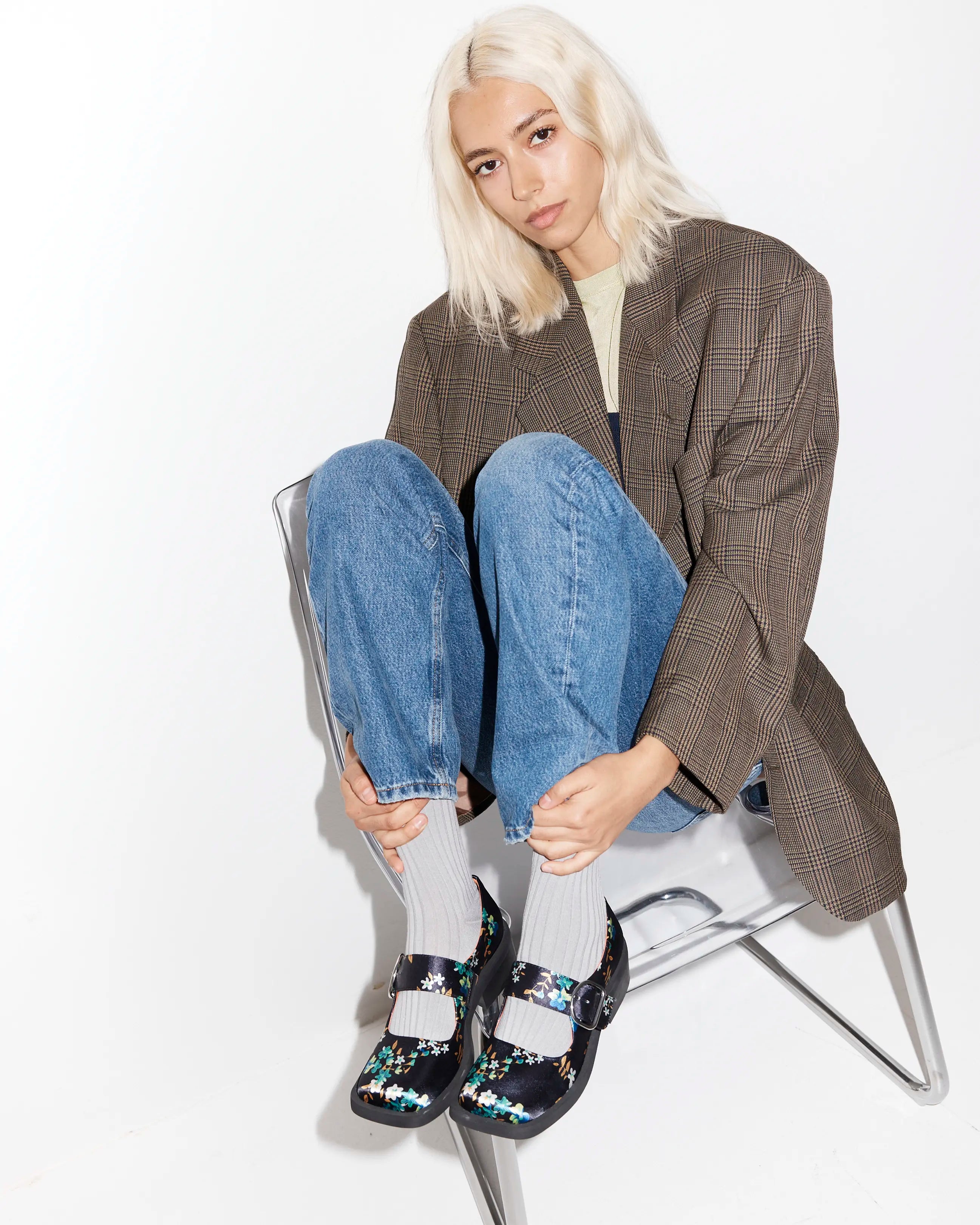Person wearing a checkered blazer, jeans, and floral patterned shoes sitting on a chair.