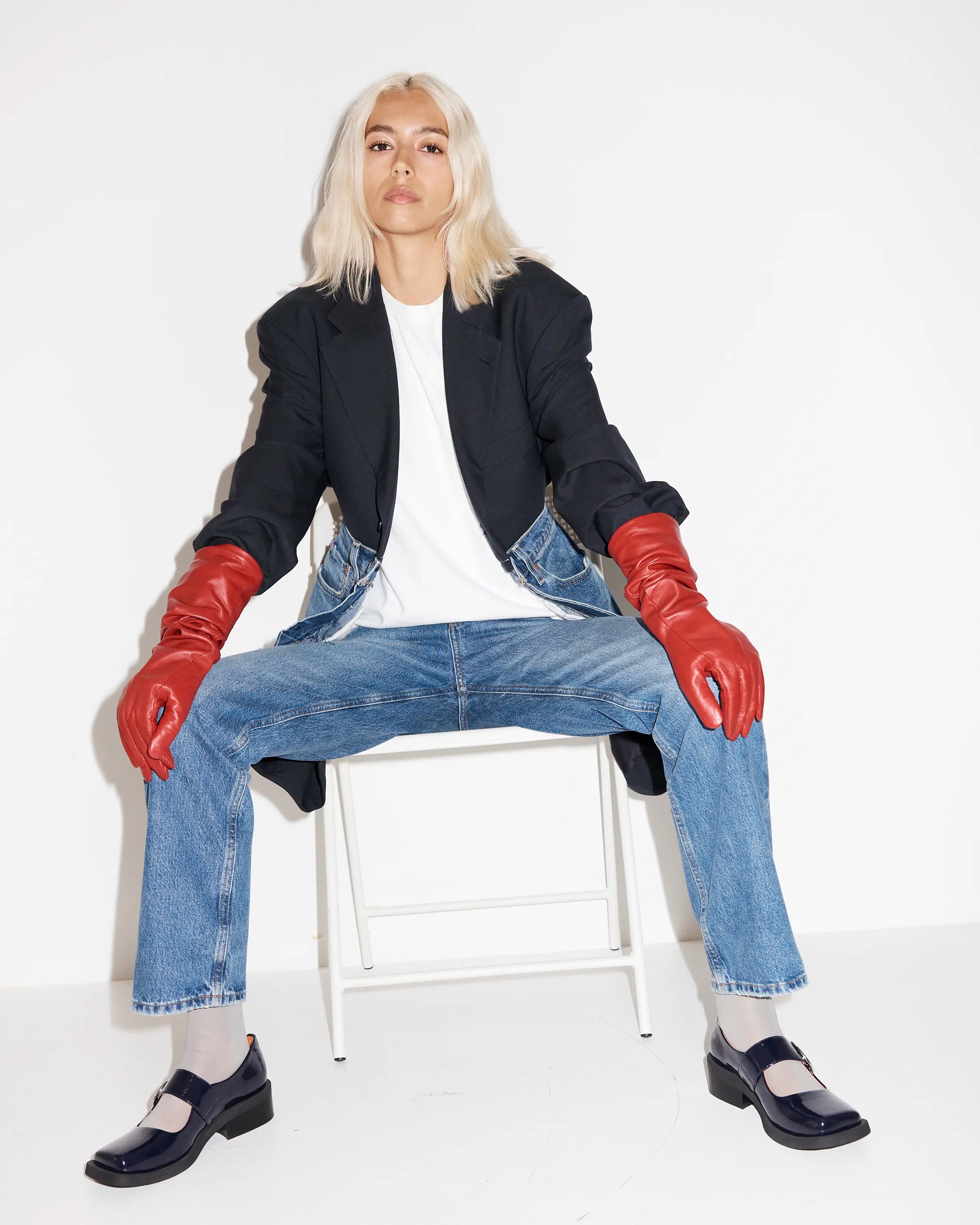 Seated person wearing a black jacket, navy square toe shoes, blue jeans, and red gloves.