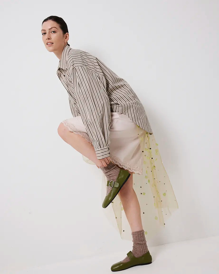 Person wearing a striped shirt, beige skirt, and green leather flat shoes with a silver buckle on a white background