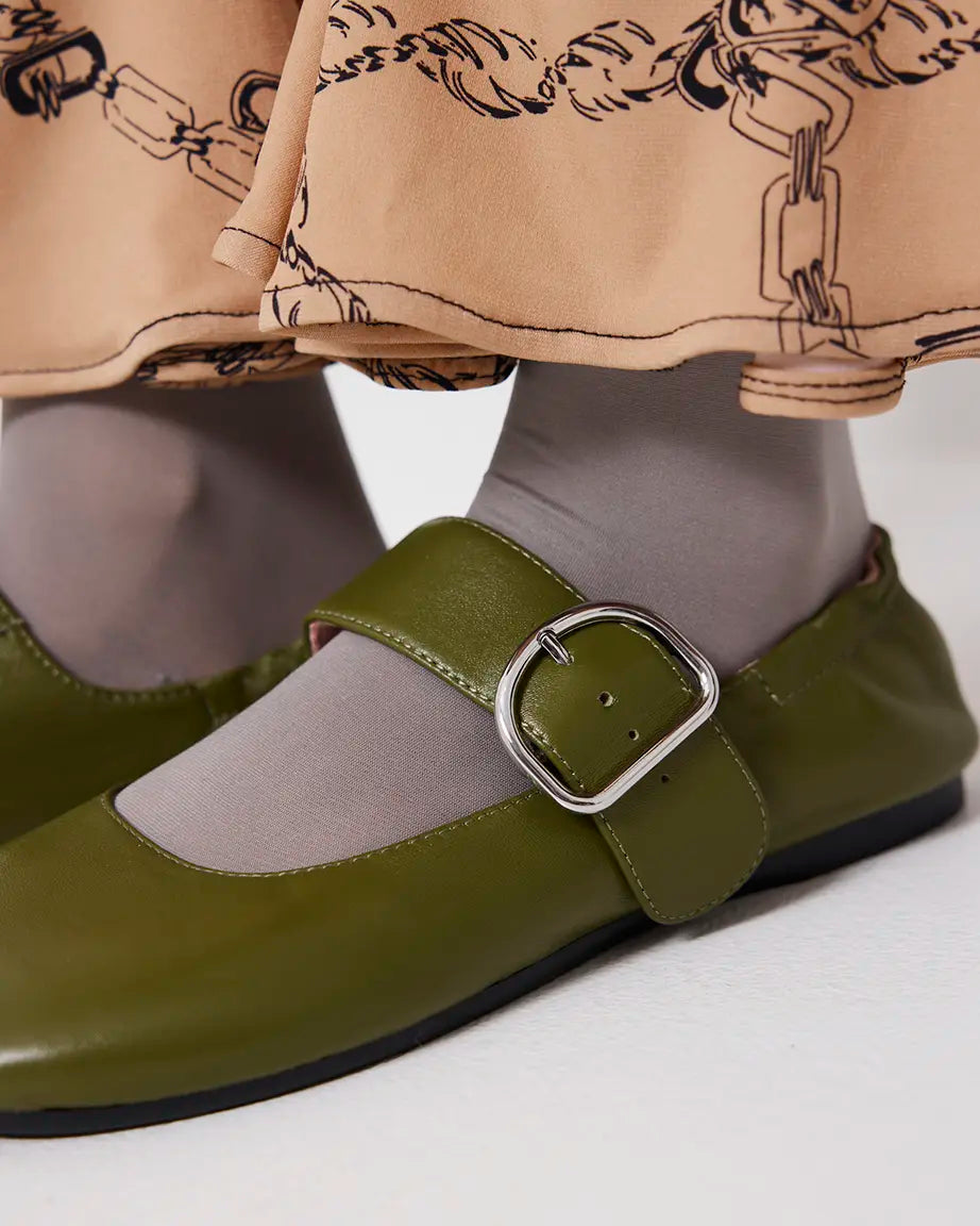 Green leather flat shoes with a silver buckle worn with beige pants and gray tights on a white background
