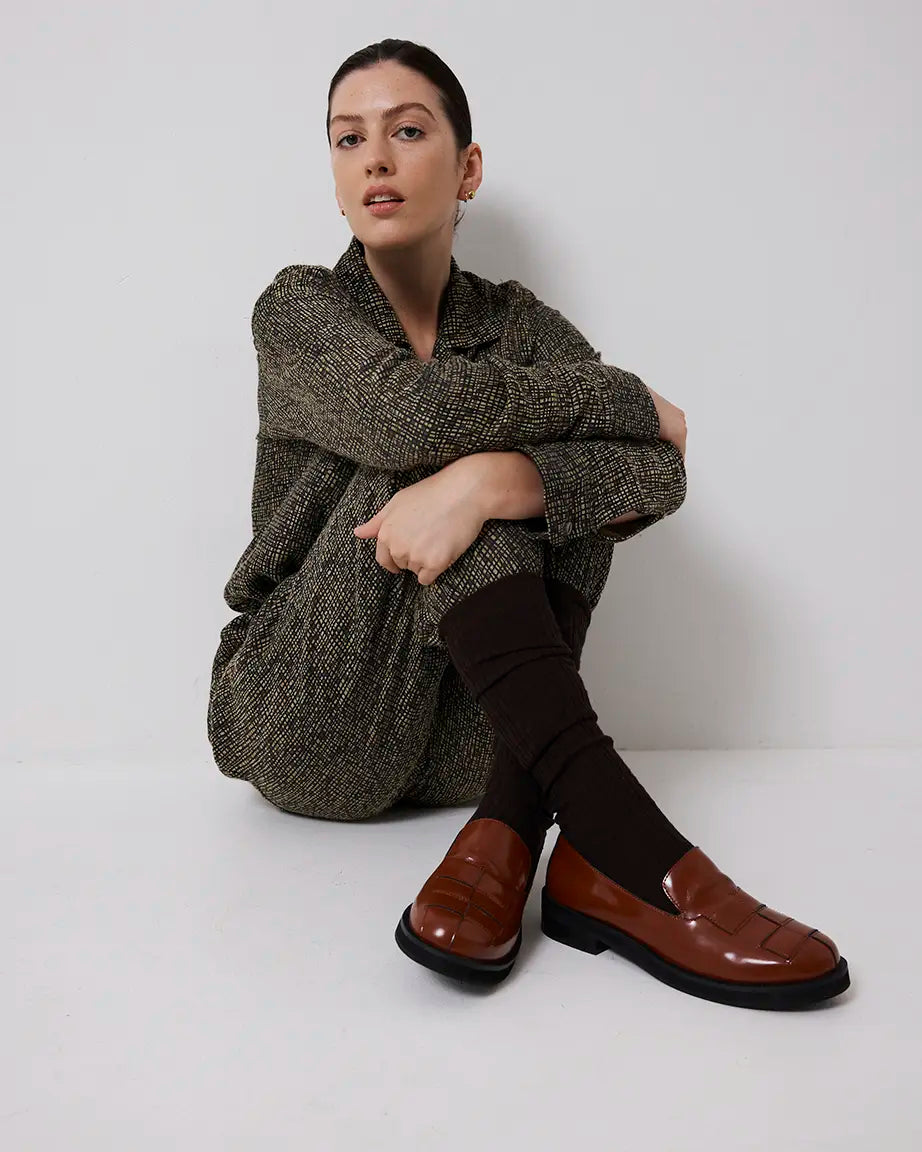 Woman wearing a brown sweater and brown leather loafers sitting on a white floor.