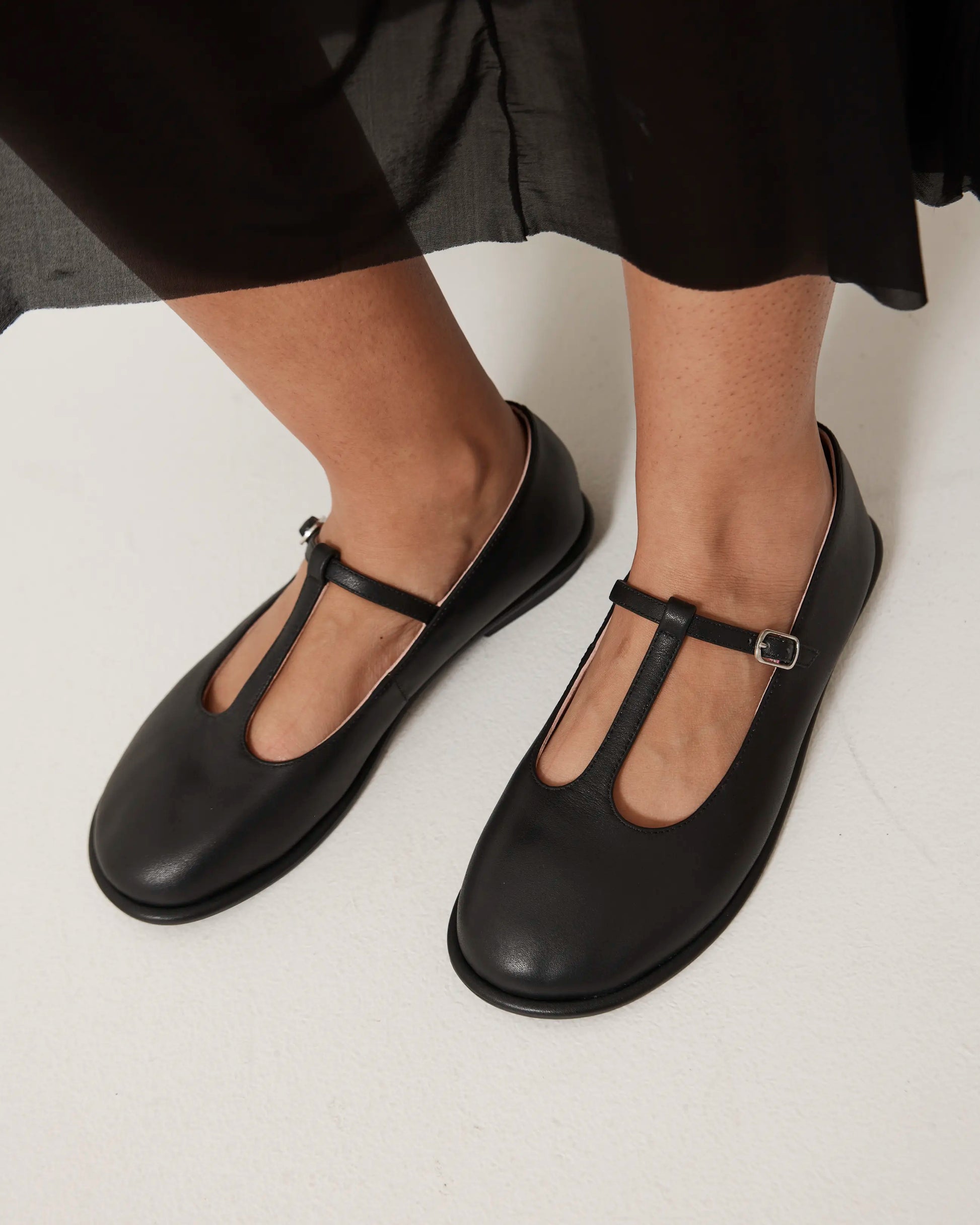 Black flat shoes with a T-strap worn by a person on a white background