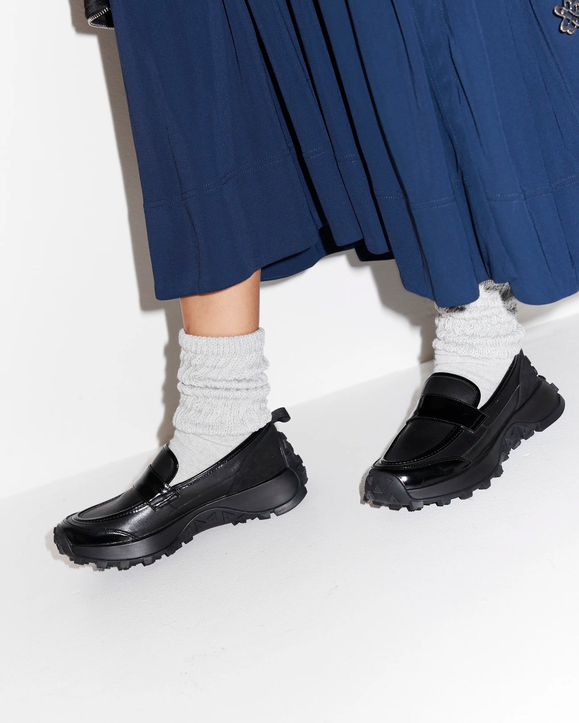 Black slip on sporty loafers worn with white socks and a blue skirt on a white background