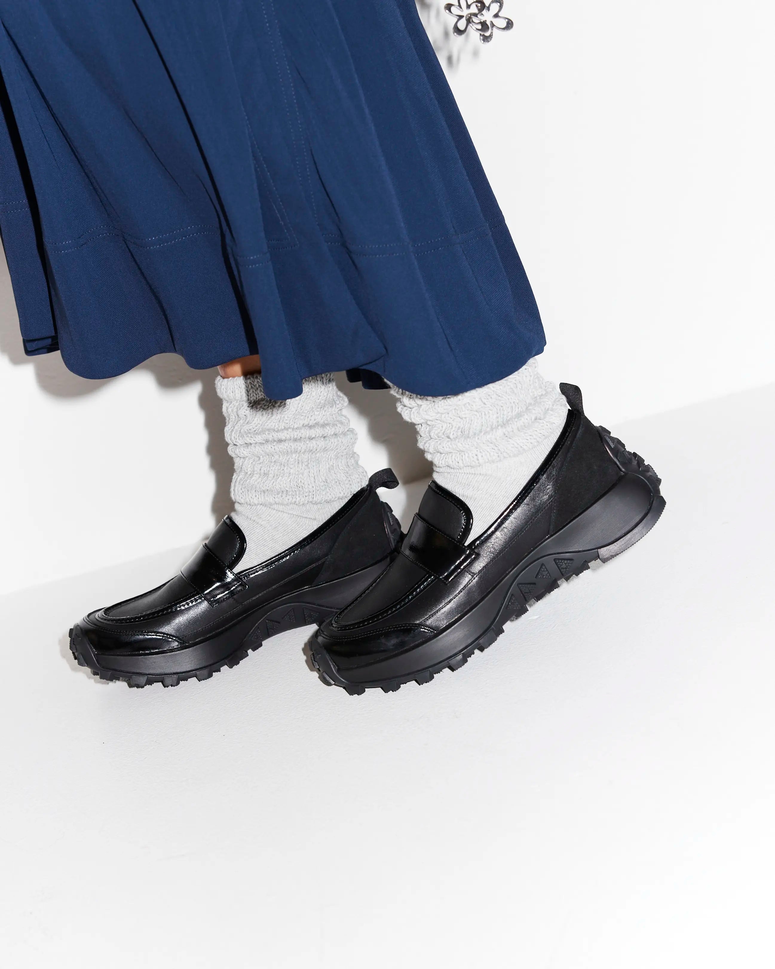 Black slip-on sporty loafers worn with a blue skirt and white socks on a light background