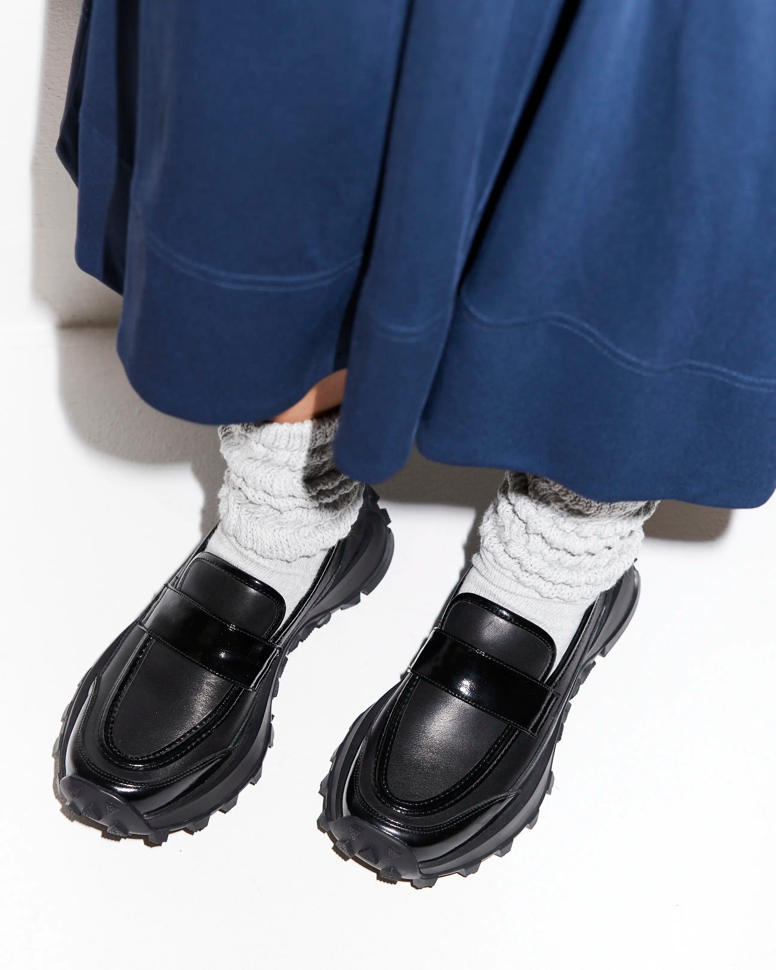 Black sporty loafers worn with white socks and a blue skirt on a white background