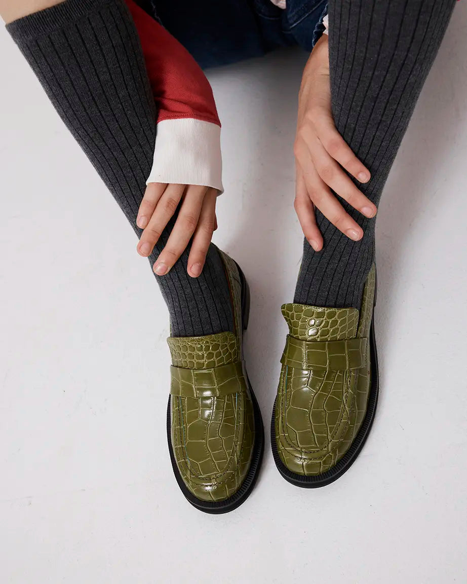 Green crocodile-textured leather loafers worn with dark ribbed socks on a white background