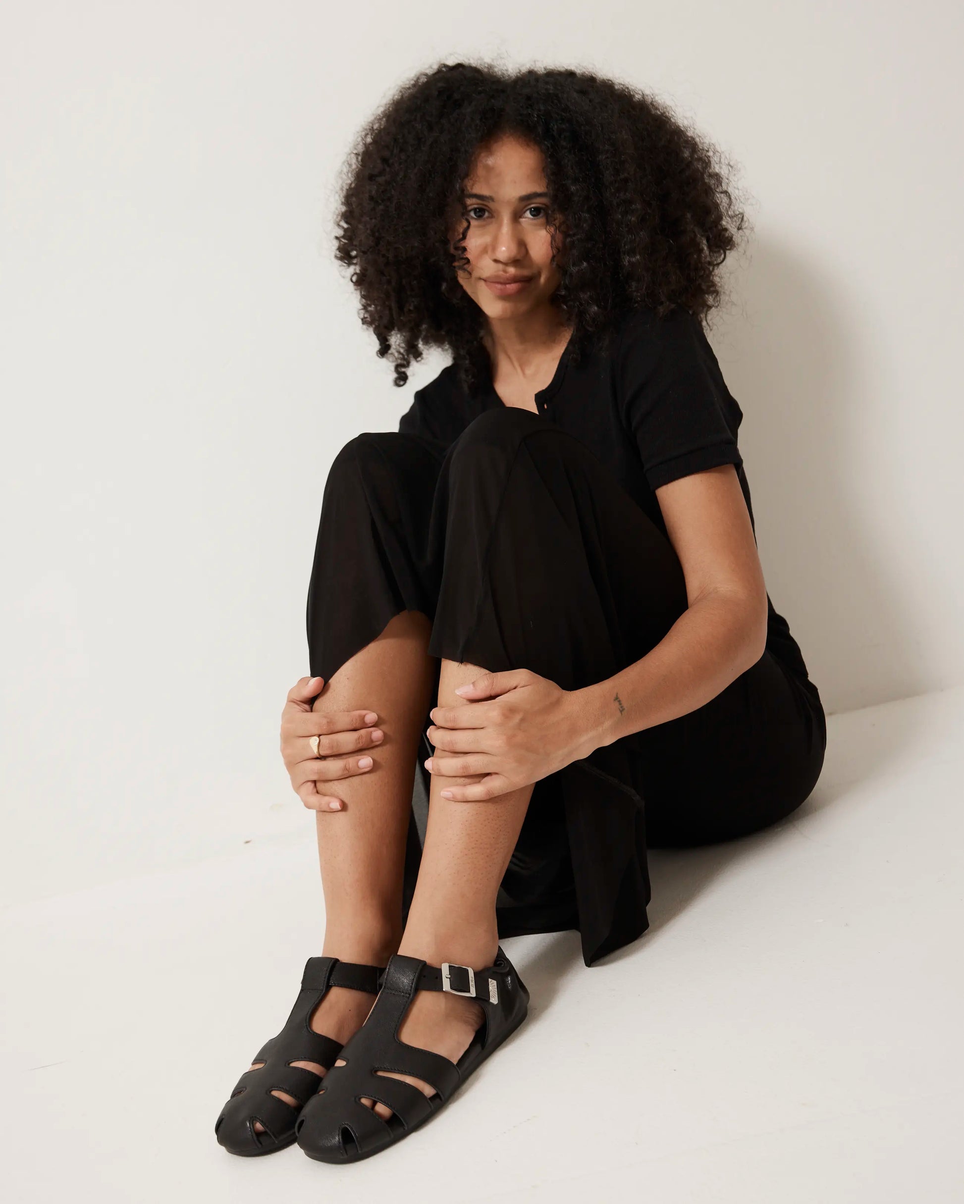Person wearing a black outfit with black fisherman sandals sitting on a white surface.