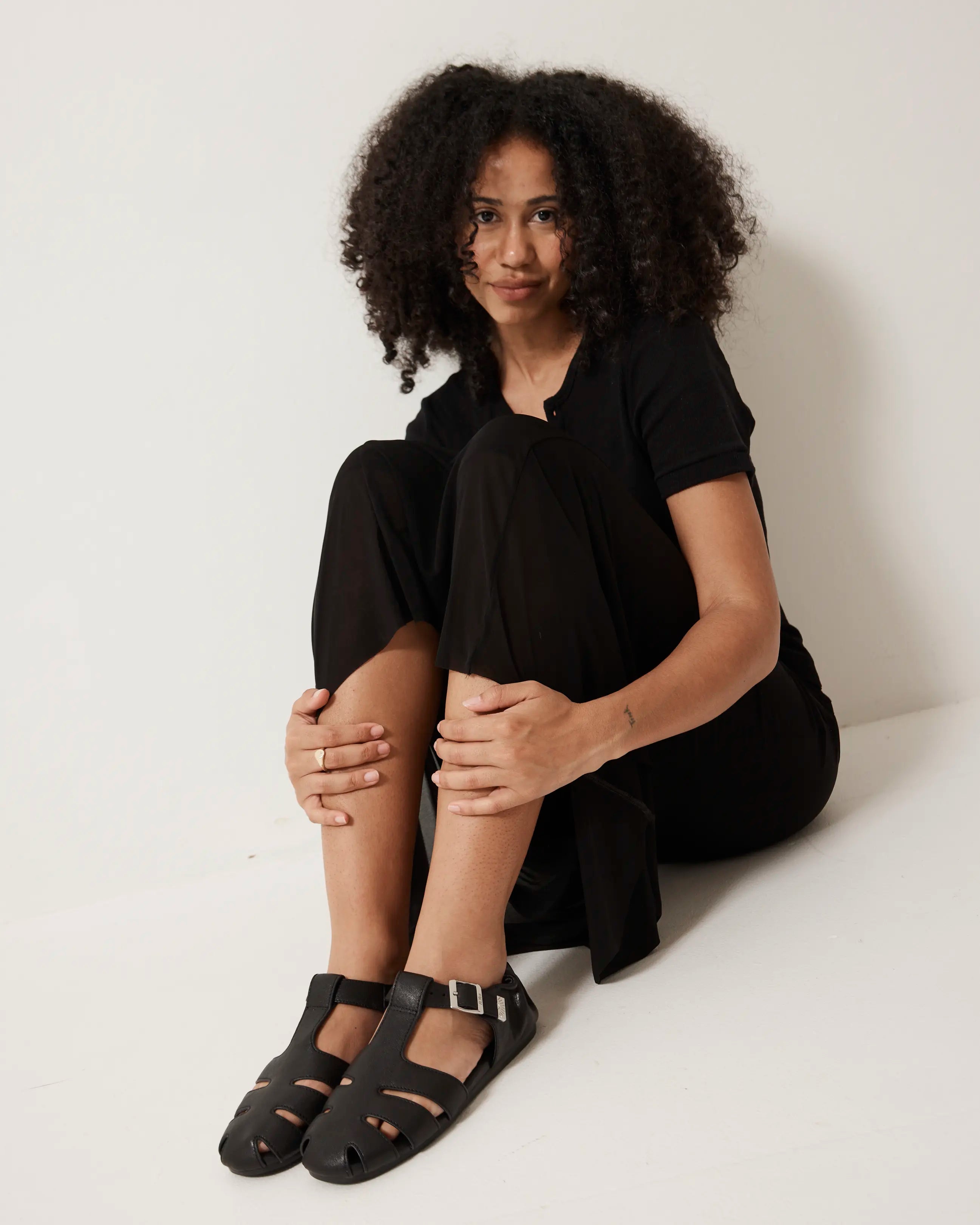 Person wearing a black outfit with black fisherman sandals sitting on a white surface.