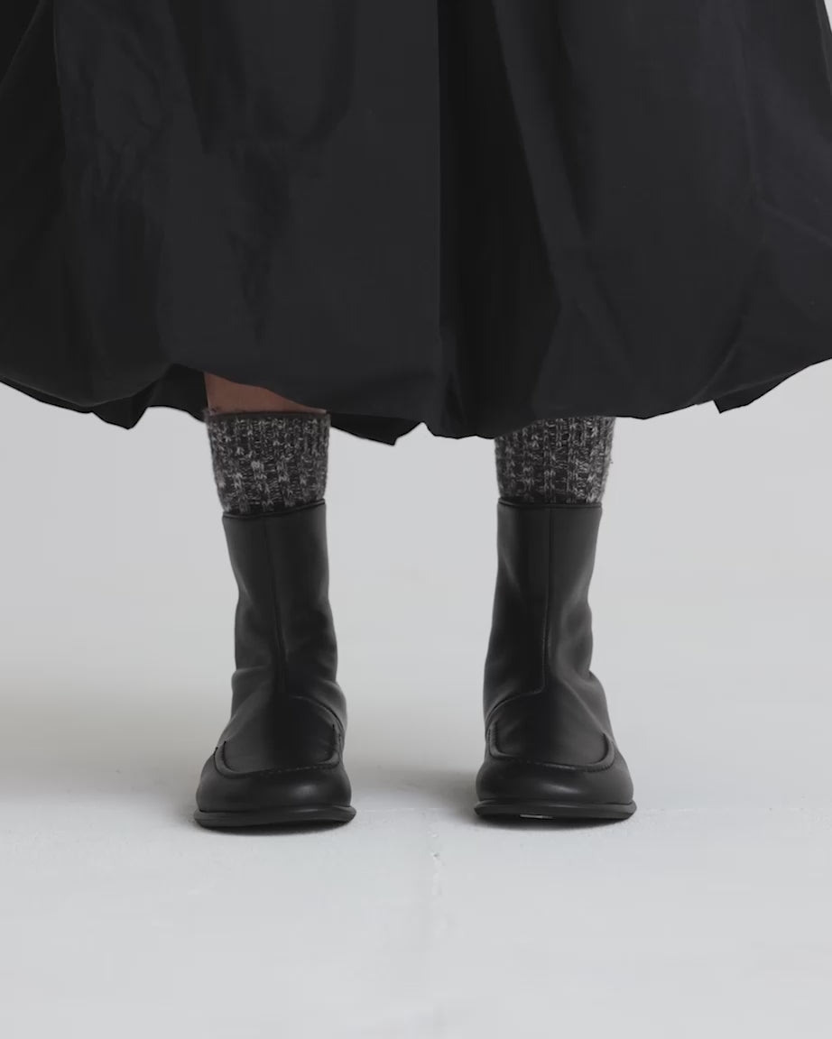 Load video: Person wearing black leather ankle boots with patterned socks and black skirt against a white background.