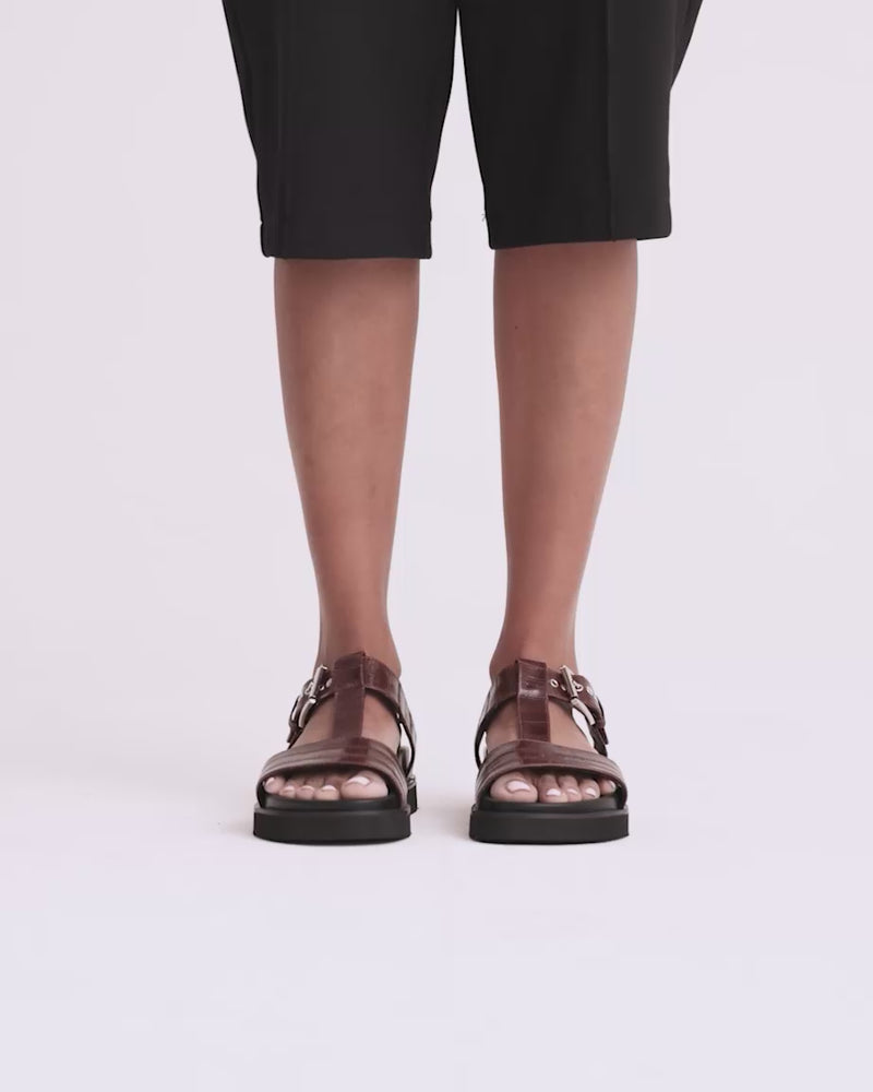 A person wearing brown leather T-bar sandals with silver buckles and arch support soles.
