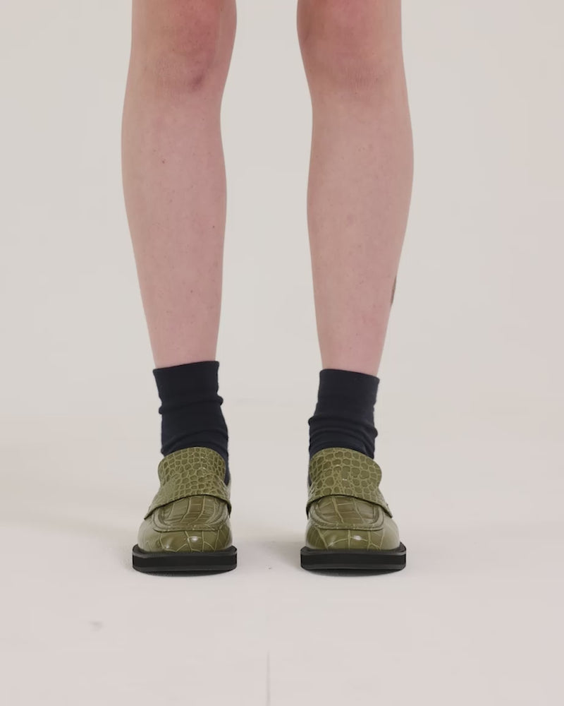 Person wearing green crocodile-textured leather loafers with black socks against a white background