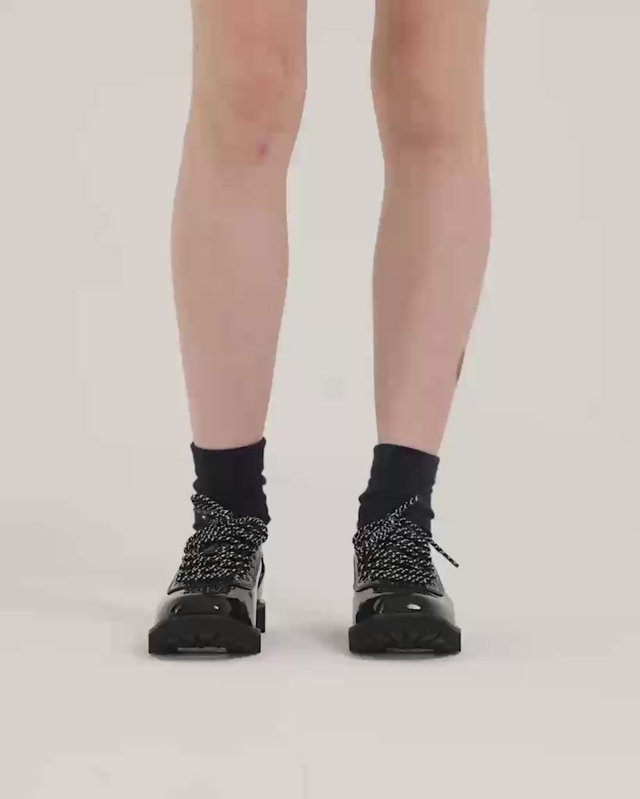 Load video: Person wearing black patent leather shoes with speckled laces and black socks.