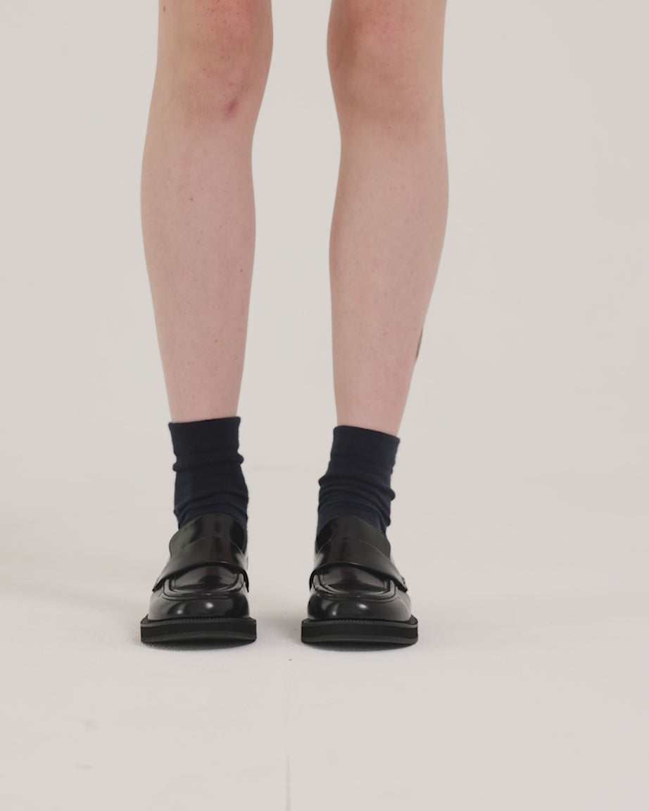 Load video: Person wearing black leather loafers with black socks against a light background.