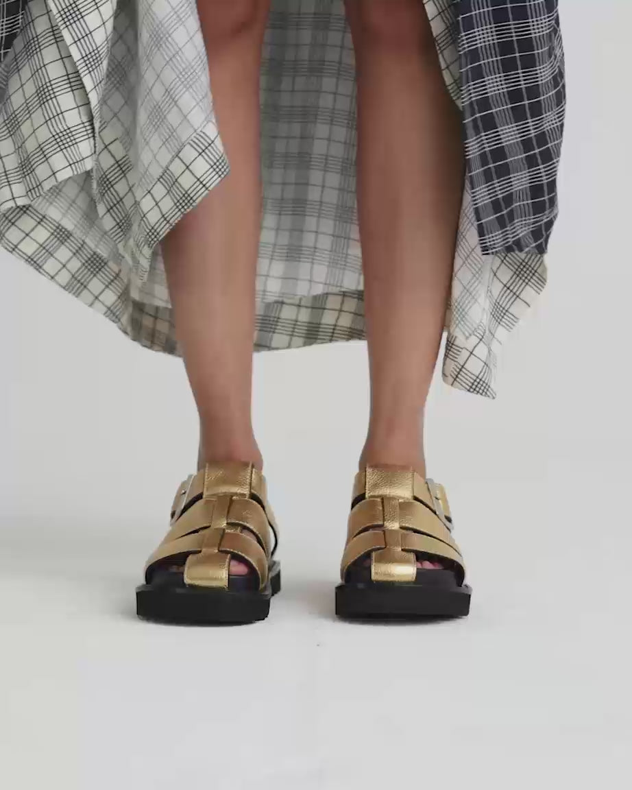 Load video: Person wearing metallic gold sandals with a black sole.